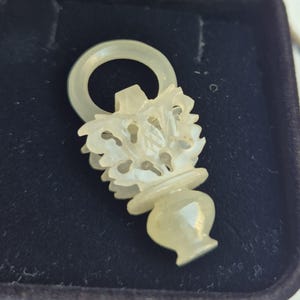 May include: A pale green, translucent carved stone pendant featuring a vase-shaped base with intricate floral detailing and a circular ring at the top. The pendant is displayed inside a black, velvet-lined jewelry box with a hinged lid.