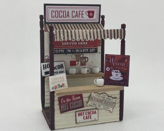 May include: A miniature Cocoa Cafe stand with a striped awning and signs that read "Cocoa Cafe", "Hot Cocoa", and "It's the Most Wonderful Time of Year". The stand features small white mugs and a sign advertising "Hot Cocoa".
