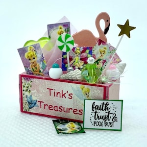 May include: A pink wooden box with the words "Tink's Treasures" on the front. The box is filled with various items, including a pink flamingo, a green lollipop, a star wand, and several fairy-themed decorations. A green card with the text "faith...trust & pixie dust" is in the foreground.