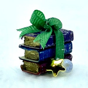 May include: A stack of miniature books with blue, red, and gold covers, tied with a green ribbon. A gold star charm and a small, light blue sphere are attached to the ribbon, creating a festive, decorative look.