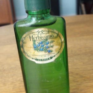May include: A rectangular green glass bottle with a dark green cap. The label features the text Herbissimo, Cologne Refresher, and Juniper, along with an illustration of blue juniper berries and green needles. The label also states 1/2 FL OZ 78.