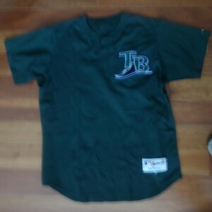 May include: A dark green short-sleeve jersey featuring the letters TB in a stylized white and light blue font on the upper left chest. A rectangular white patch with red and blue text is stitched onto the lower left front of the garment.