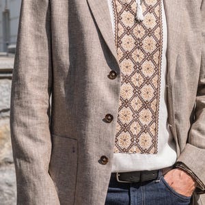 May include: A man wearing a light brown linen blazer over a white embroidered shirt with a brown and beige pattern. He is wearing dark blue jeans and a black belt.
