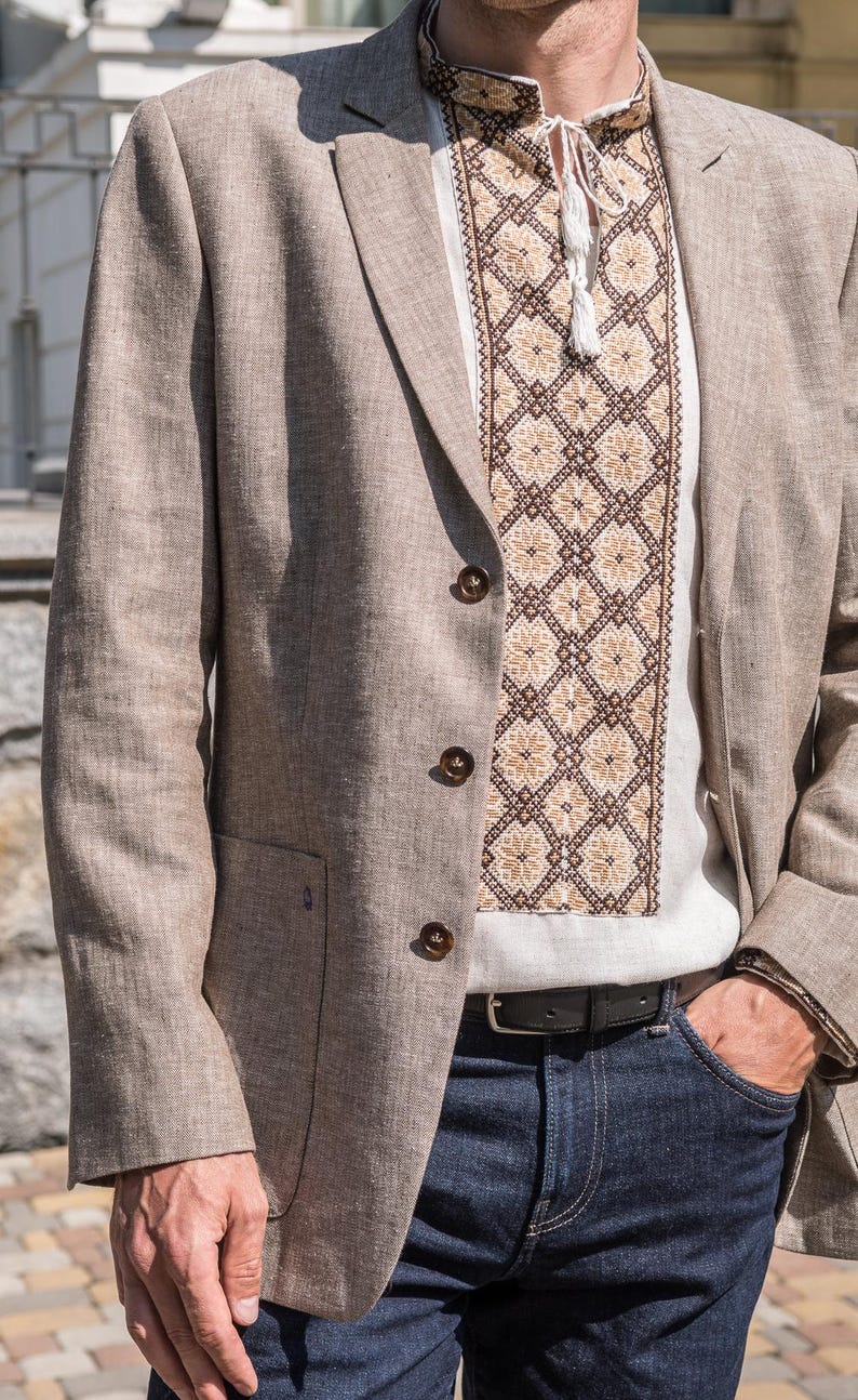 May include: A man wearing a light brown linen blazer over a white embroidered shirt with a brown and beige pattern. He is wearing dark blue jeans and a black belt.