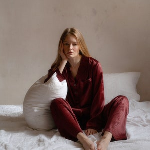 May include: A person wearing a red and black checked pyjama set sits on a white bed with a white pillow. They are looking at the camera with a thoughtful expression.