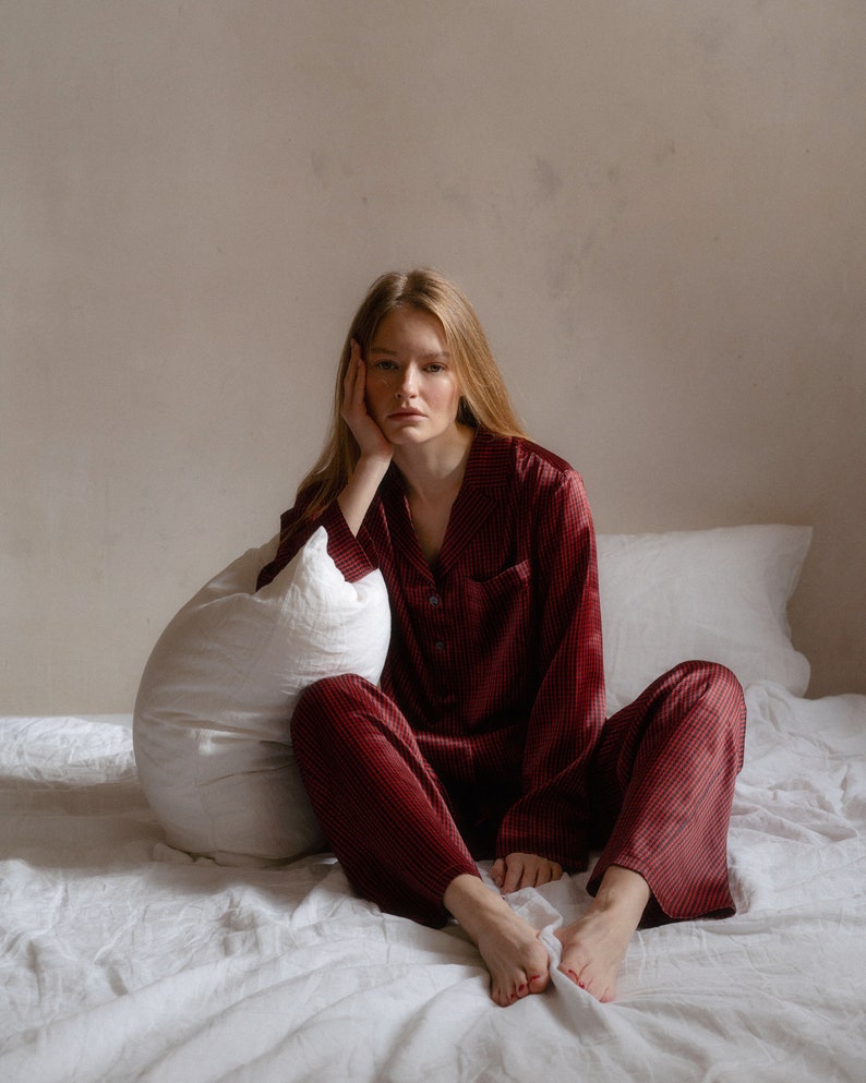 May include: A person wearing a red and black checked pyjama set sits on a white bed with a white pillow. They are looking at the camera with a thoughtful expression.