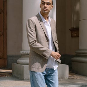 May include: A man wearing a light brown linen blazer over a white button-down shirt and blue jeans.