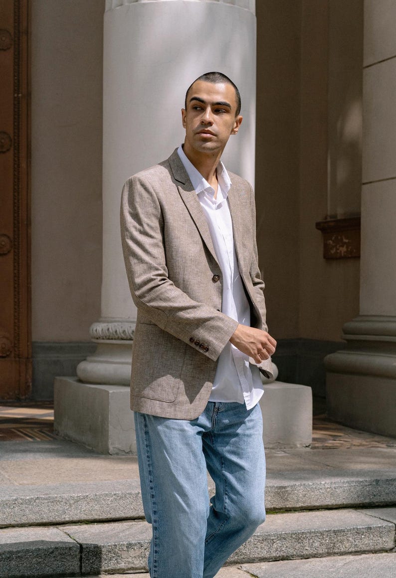 May include: A man wearing a light brown linen blazer over a white button-down shirt and blue jeans.
