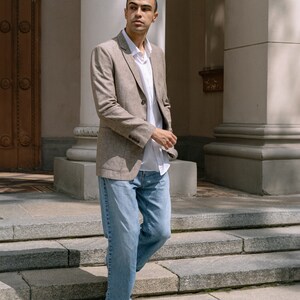 May include: A man wearing a light brown blazer over a white button-down shirt and blue jeans. He is standing on a set of stone steps.