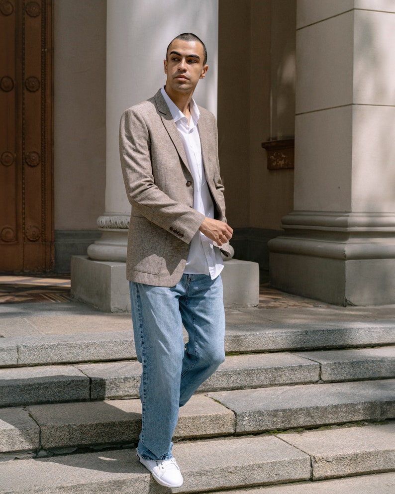 May include: A man wearing a light brown blazer over a white button-down shirt and blue jeans. He is standing on a set of stone steps.