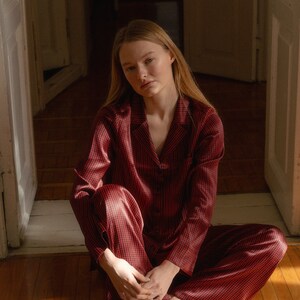 May include: A woman sits on a wooden floor wearing a red and black checked pyjama set. The pyjamas have a button-up top and long trousers.