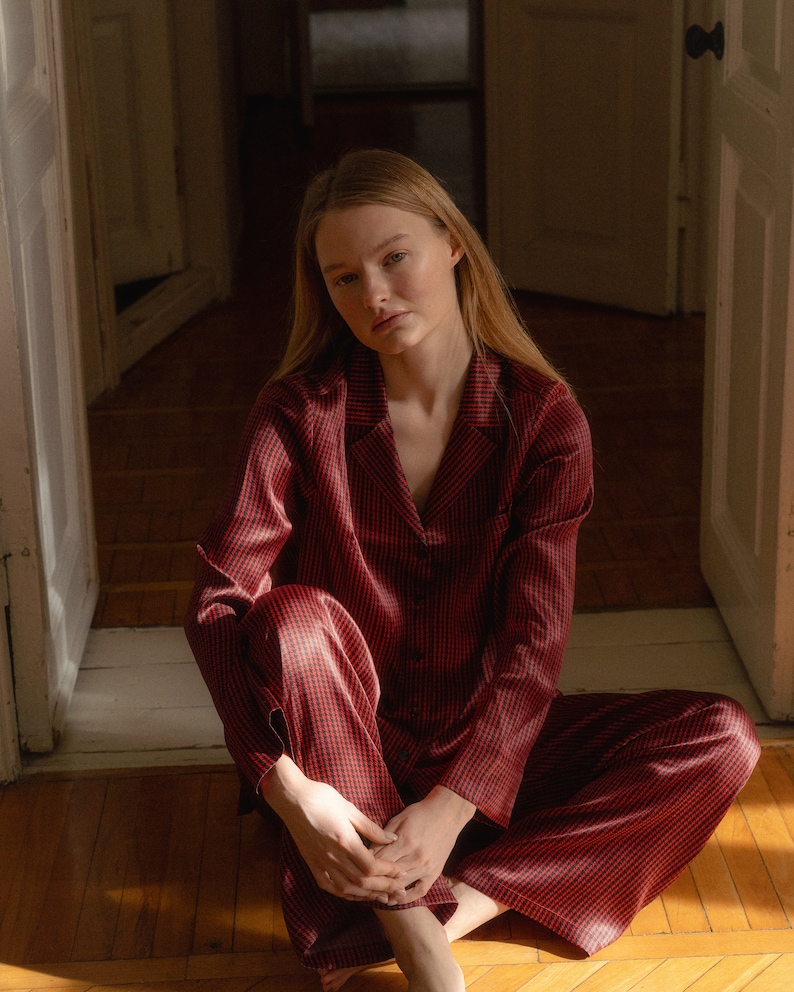 May include: A woman sits on a wooden floor wearing a red and black checked pyjama set. The pyjamas have a button-up top and long trousers.