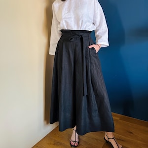 May include: A black linen skirt with a wide black fabric belt. The skirt is long and flowing, and the belt is tied in a bow at the front. The skirt is worn with a white linen top.