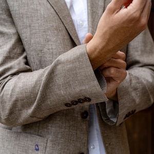 May include: A brown linen blazer with a white button-down shirt underneath. The blazer has a pocket on the left side and buttons on the front. The person is adjusting the cuff of the blazer.