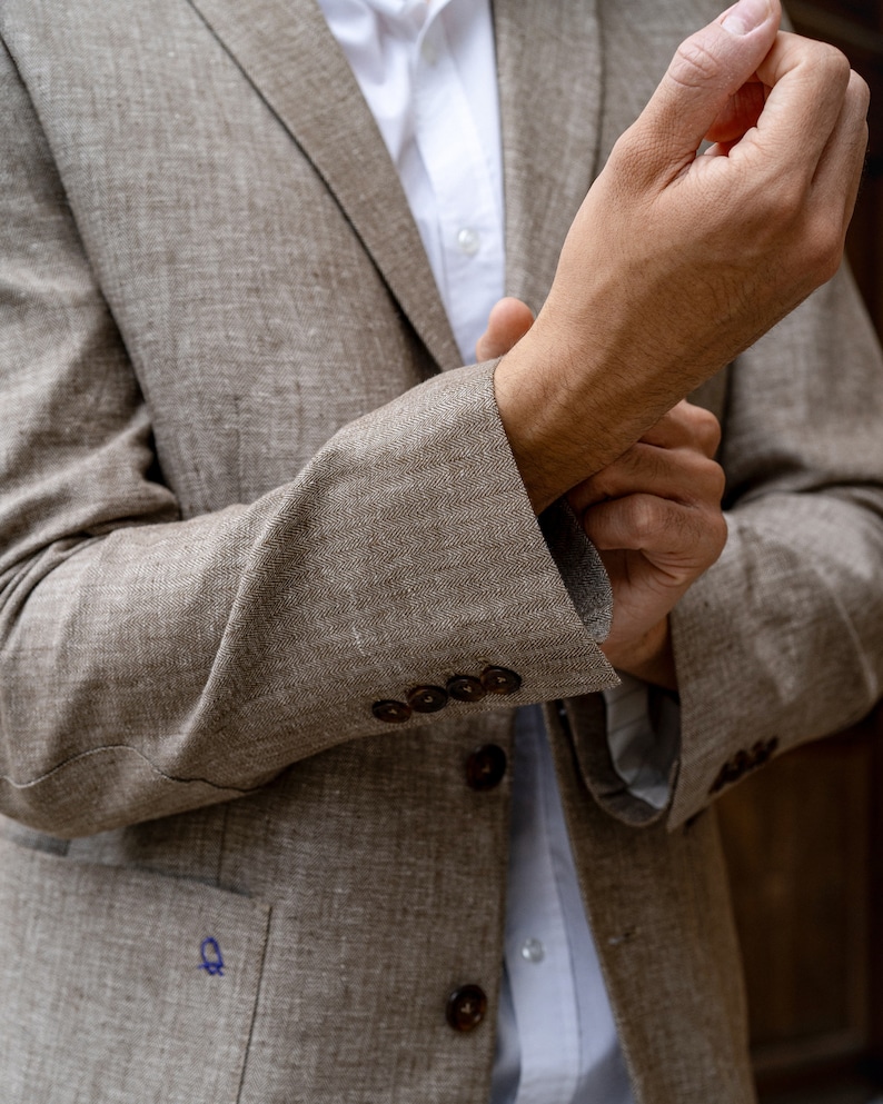 May include: A brown linen blazer with a white button-down shirt underneath. The blazer has a pocket on the left side and buttons on the front. The person is adjusting the cuff of the blazer.