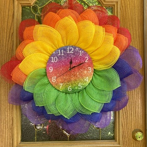 May include: A rainbow-colored mesh wreath with a glitter clock in the center. The clock face has white numbers and hands. The wreath is decorated with a red ribbon.