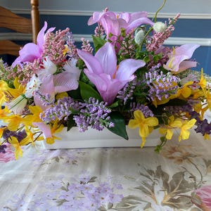 May include: A floral arrangement in a white rectangular container. The arrangement features pink lilies, yellow daffodils, purple lavender, and other colorful flowers. The flowers are arranged on a floral patterned tablecloth.