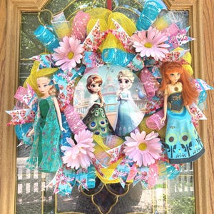 Large 22-inch Frozen Fever wreath featuring 11.5-inch Elsa and Anna dolls in summer floral dresses with a central metal sign on a decorative ribbon base.