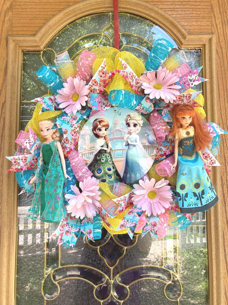 Large 22-inch Frozen Fever wreath featuring 11.5-inch Elsa and Anna dolls in summer floral dresses with a central metal sign on a decorative ribbon base.