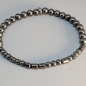 May include: A dark gray beaded bracelet. The bracelet features a mix of round and cylindrical beads. The beads are strung together, forming a flexible band. The bracelet is designed to be worn around the wrist.