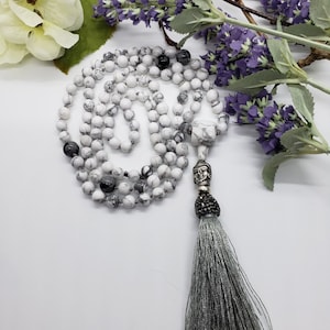 May include: White and gray beaded mala necklace with a silver Buddha head pendant and a gray tassel. The necklace is made of round, marbled beads. Lavender and white flowers are in the background.