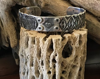 May include: A silver cuff bracelet with a geometric pattern and a heart design. The bracelet is on a display stand made of wood.