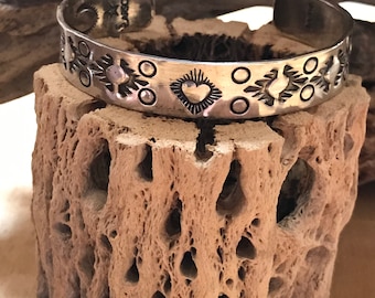 May include: A silver cuff bracelet with a heart design and geometric patterns. The bracelet is stamped with the word "LARSON".