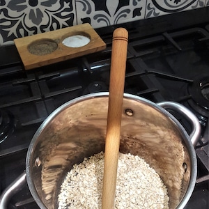 Peut inclure: Une casserole en acier inoxydable avec une cuillère en bois à l'intérieur. La casserole est remplie d'avoine en flocons non cuits. La casserole est sur une cuisinière noire.