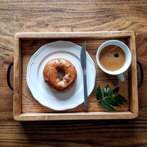 May include: A wooden tray with a white plate, a cup of coffee, and a bagel. The tray is on a wooden table. There is a sprig of green leaves on the tray.
