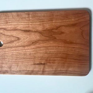 Op de afbeelding: Een rechthoekig snijplank van kersenhout met een gat in de linkerbovenhoek. De plank heeft een natuurlijke houtkleur en een glad oppervlak.