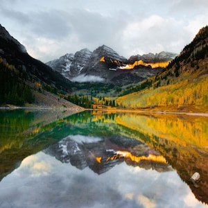May include: A mountain lake with a reflection of the surrounding mountains and trees. The trees are a mix of green and yellow, indicating autumn. The water is still and clear, reflecting the sky and clouds.