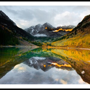 May include: A scenic view of a mountain lake with a reflection of the surrounding mountains and trees. The lake is surrounded by trees with yellow and orange leaves, and the mountains are covered in snow. The sky is cloudy and the water is still.