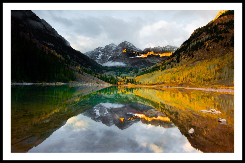 May include: A scenic view of a mountain lake with a reflection of the surrounding mountains and trees. The lake is surrounded by trees with yellow and orange leaves, and the mountains are covered in snow. The sky is cloudy and the water is still.