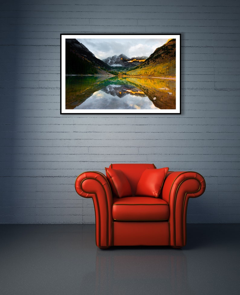 May include: A red armchair with a single pillow sits in front of a brick wall with a framed photograph of a mountain lake reflecting the surrounding mountains.