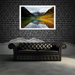 May include: A framed photograph of a mountain lake with fall foliage reflected in the water. The lake is surrounded by mountains with snow-capped peaks. The sky is a light blue with some clouds.