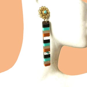 May include: A gold-colored stud earring with a turquoise stone center and a decorative filigree design. The earring hangs a long, rectangular pendant with alternating stripes of black, turquoise, orange, white, and silver. Jewelry.