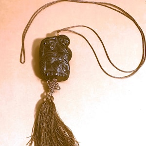 May include: A dark brown wooden pendant with a carved image of three monkeys. The pendant is attached to a brown cord with a tassel.
