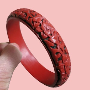May include: A red carved bracelet with floral designs. The bracelet is a solid red color with intricate carvings of flowers and leaves. The background is a light pink color.