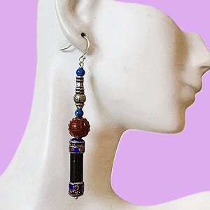 May include: A long, dangling earring with a silver hook. The earring features a combination of beads, including blue, silver, and a carved reddish-brown bead. The bottom portion is a black cylinder with blue and red accents. The earring is displayed against a light purple background.