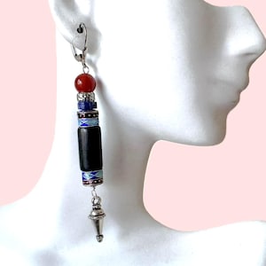 May include: A long, dangling earring with a silver leverback clasp. The earring features a red bead, blue and silver beads, a black cylindrical bead, and a silver cone-shaped charm. The beads have intricate designs.