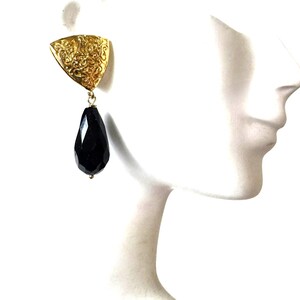 May include: A gold-colored earring with a triangular top featuring an ornate design and a black, teardrop-shaped gemstone pendant. The earring is displayed on a white mannequin head, showcasing its design and shape.