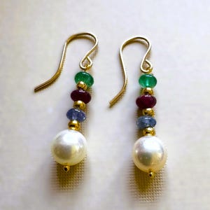 May include: A pair of gold-colored earrings with a hook design. Each earring features a sequence of green, red, and blue gemstones, with a large white pearl at the bottom. The earrings are designed for pierced ears.