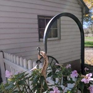 May include: A brown giraffe-shaped plant stake is used to support a potted plant with pink flowers. The plant is in front of a white picket fence and a white house with a black metal arch.