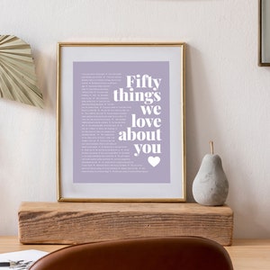 May include: A framed print with a light purple background and white text that reads "Fifty things we love about you" with a small white heart at the bottom. The frame is gold and the print is sitting on a wooden shelf.