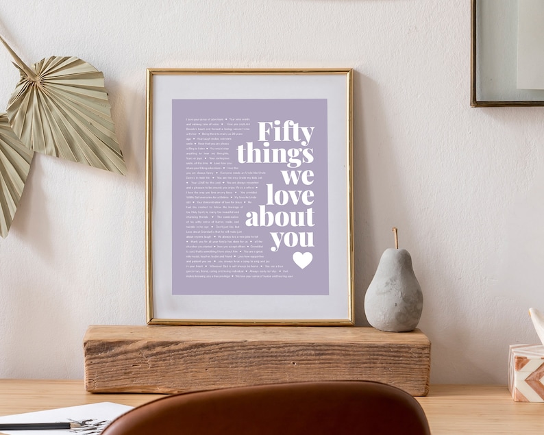 May include: A framed print with a light purple background and white text that reads "Fifty things we love about you" with a small white heart at the bottom. The frame is gold and the print is sitting on a wooden shelf.