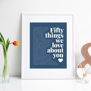 May include: A framed print with a blue background and white text that reads "Fifty things we love about you" with a small heart at the bottom. The print is in a white frame and is sitting on a white surface.