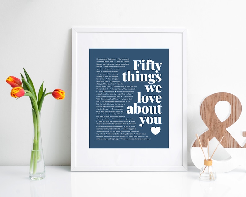 May include: A framed print with a blue background and white text that reads "Fifty things we love about you" with a small heart at the bottom. The print is in a white frame and is sitting on a white surface.