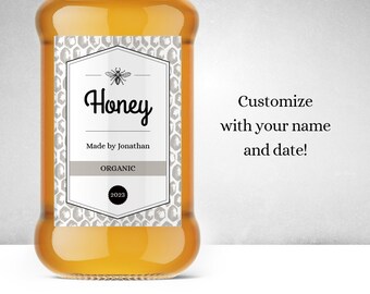 Könnte beinhalten: Ein Glas Honig mit einem weißen Etikett. Das Etikett hat ein Wabenmuster und sagt "Honey" in schwarzer Kursivschrift. Es steht auch "Made by Jonathan" und "Organic" darauf. Das Jahr "2023" ist in einem Kreis auf dem Etikett gedruckt.