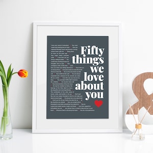 May include: A framed print with a grey background and white text that reads "Fifty things we love about you" with a red heart at the bottom. The print is in a white frame and is sitting on a white surface.