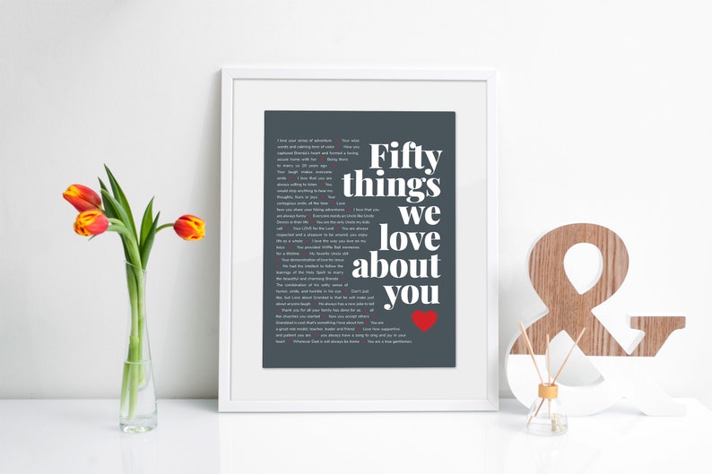 May include: A framed print with a grey background and white text that reads "Fifty things we love about you" with a red heart at the bottom. The print is in a white frame and is sitting on a white surface.